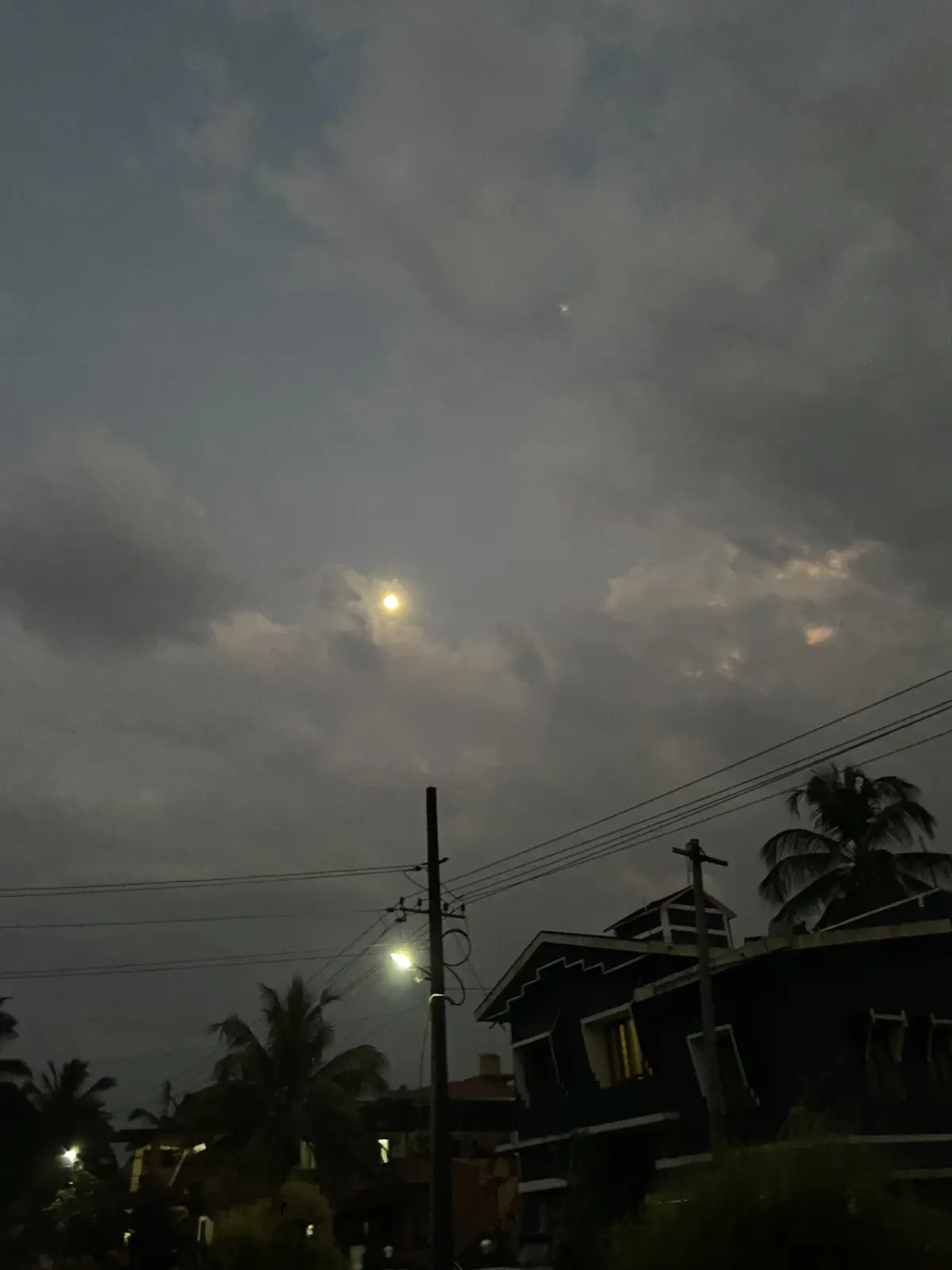A nighttime photo shows a bright moon partially obscured by clouds above a residential area with houses, palm trees, and…