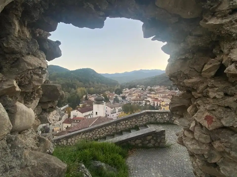 A scenic view of a European village nestled in hills, framed by a rough stone archway. The image shows a cobblestone path…