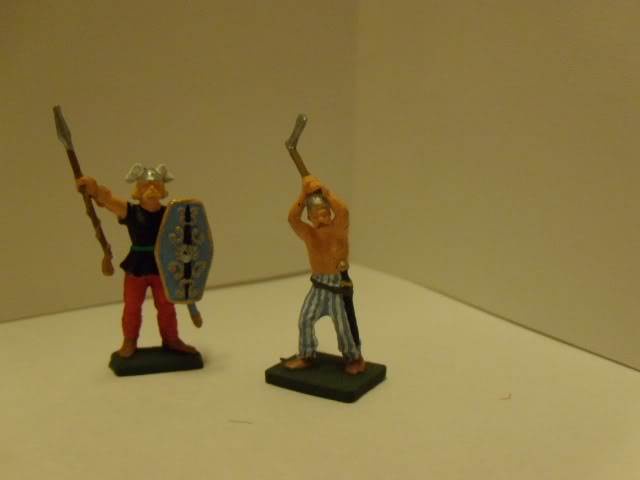Two small plastic Viking figurines stand on a white surface; one holds a spear and shield, the other holds a sword above his…