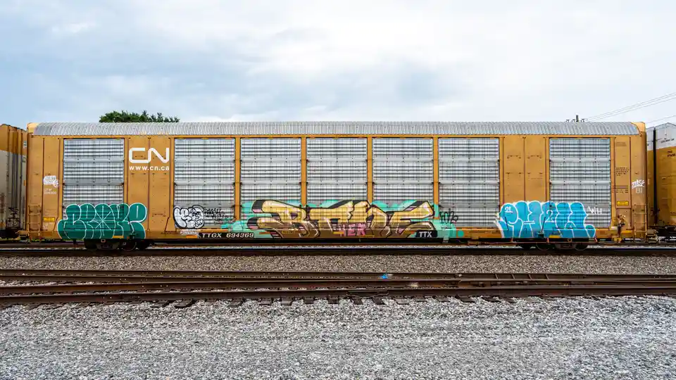 A yellow Canadian National (CN) freight train car sits on tracks under a cloudy sky, covered in colorful graffiti. The…