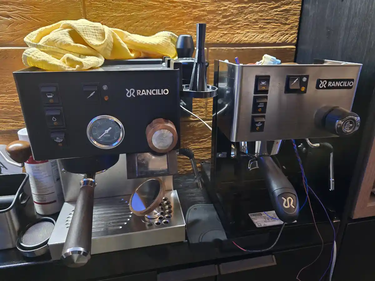 Two Rancilio espresso machines sit side by side on a counter; the left machine is black with a visible pressure gauge and a…