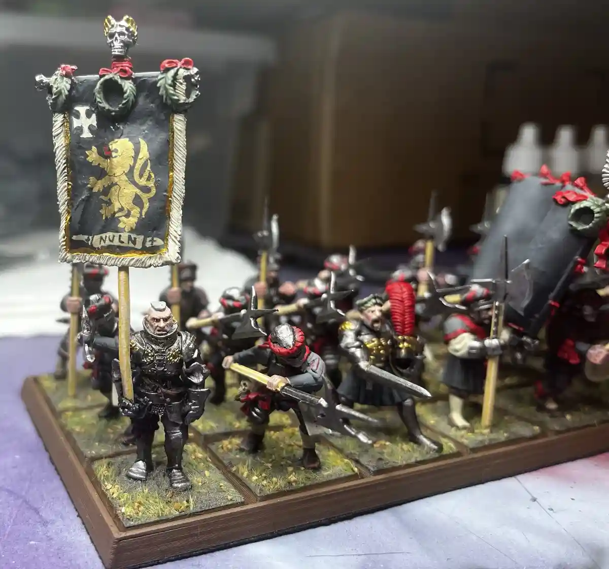 A photo of painted miniature fantasy warband figures on a wooden base, led by a heavily armored warrior holding a black…