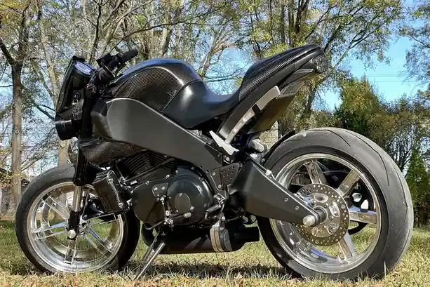A black custom motorcycle with carbon fiber accents and chrome wheels is parked on grass under trees. The overlaid text…
