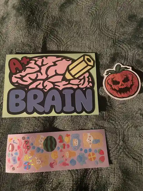 Three stickers lie on a dark gray textured surface: a large one with a pink brain, a yellow pencil, and the text 'A+ BRAIN'…