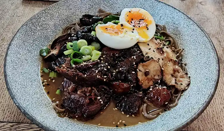 A close-up photo of a bowl of dark broth noodle soup with soba noodles, topped with sliced mushrooms, chopped green onions…