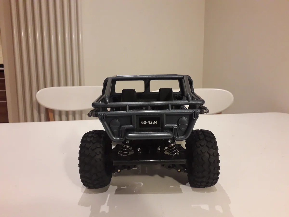 A black remote-controlled off-road vehicle with large knobby tires and an open-frame body sits on a white table, facing the…