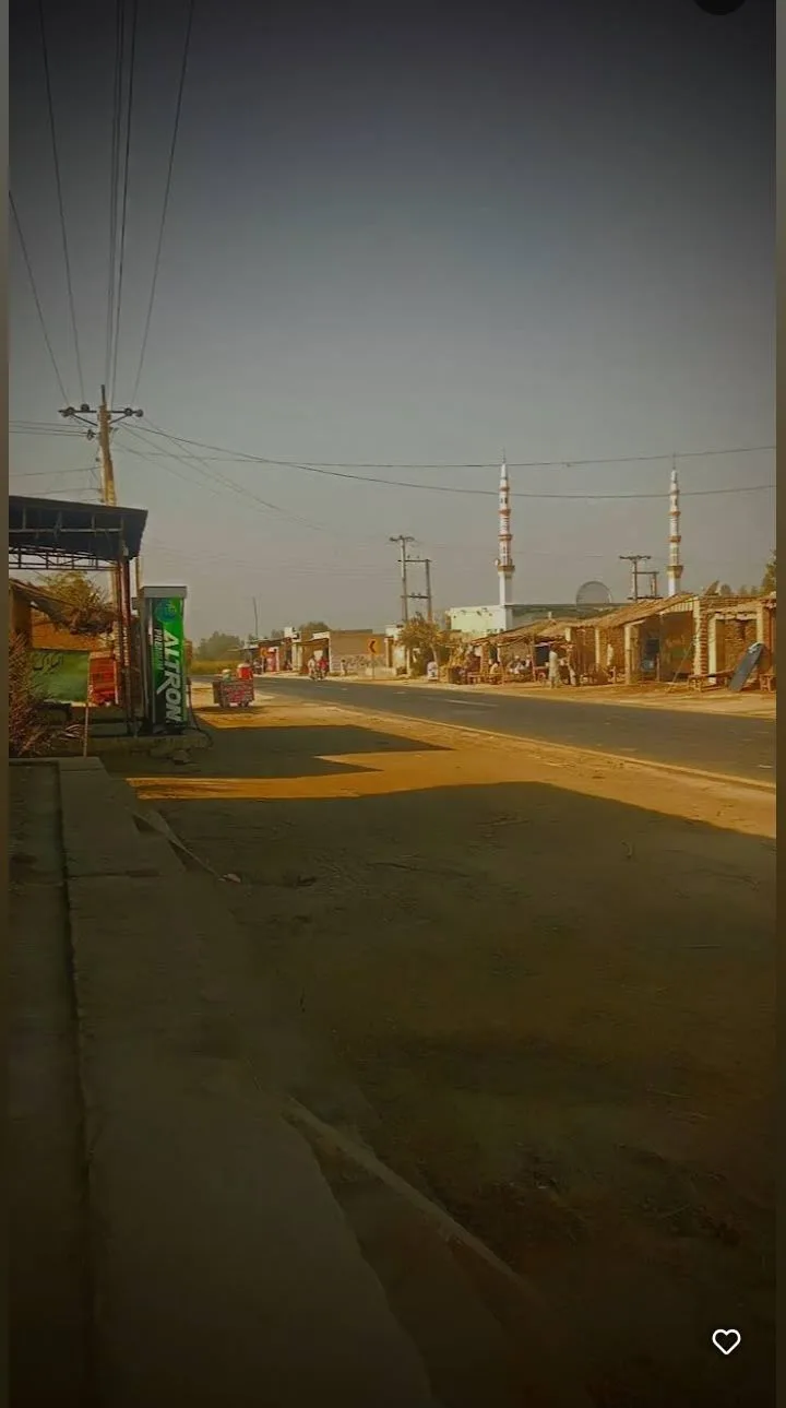 A low-angle street photo shows a quiet road with simple buildings, overhead power lines, and two mosque minarets in the…