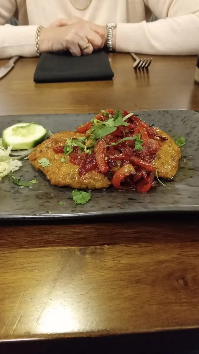 A close-up photo shows a plated dish of breaded chicken topped with red peppers and cilantro, served with cucumber and…