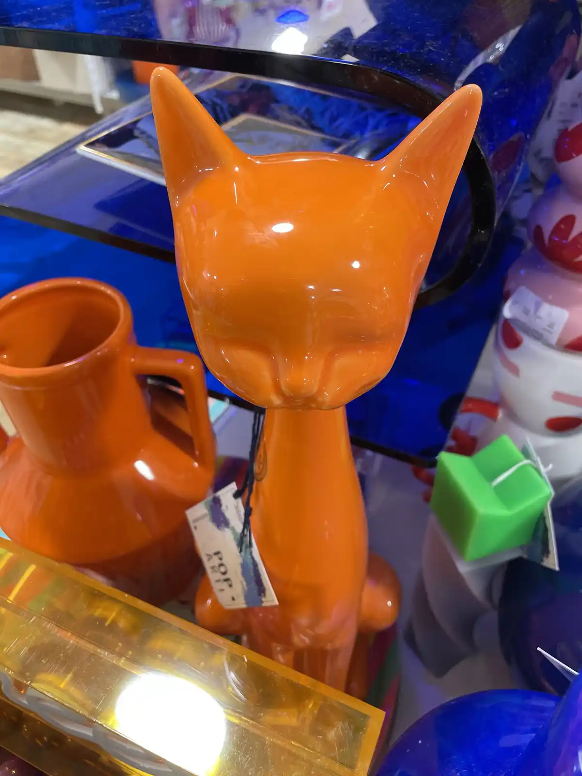 A glossy orange ceramic cat figurine with a long neck and pointed ears stands on a shelf surrounded by colorful glassware…