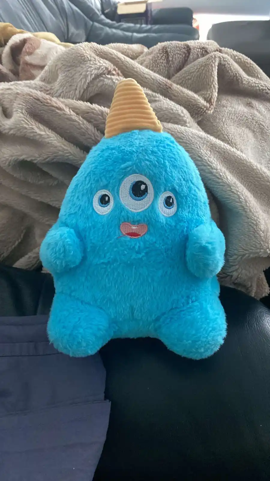 A blue, fuzzy plush toy with three embroidered eyes, a small pink mouth, and a yellow cone-shaped horn sits on a dark couch…