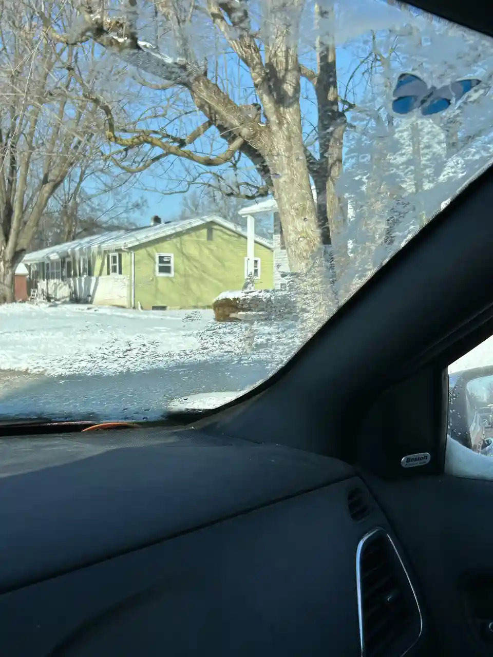 A photo taken from inside a car looking out the driver's side window, which is covered in frost and ice crystals. Outside, a…