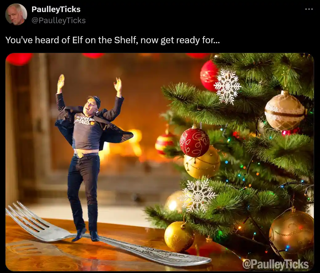A man in a Santa-themed shirt and cap stands with arms raised on a large silver fork, positioned on a wooden table next to a…
