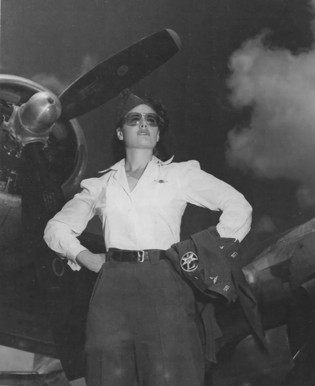 A black-and-white historical photo shows a confident woman in a U.S. Army Air Forces uniform, standing with hands on hips in…