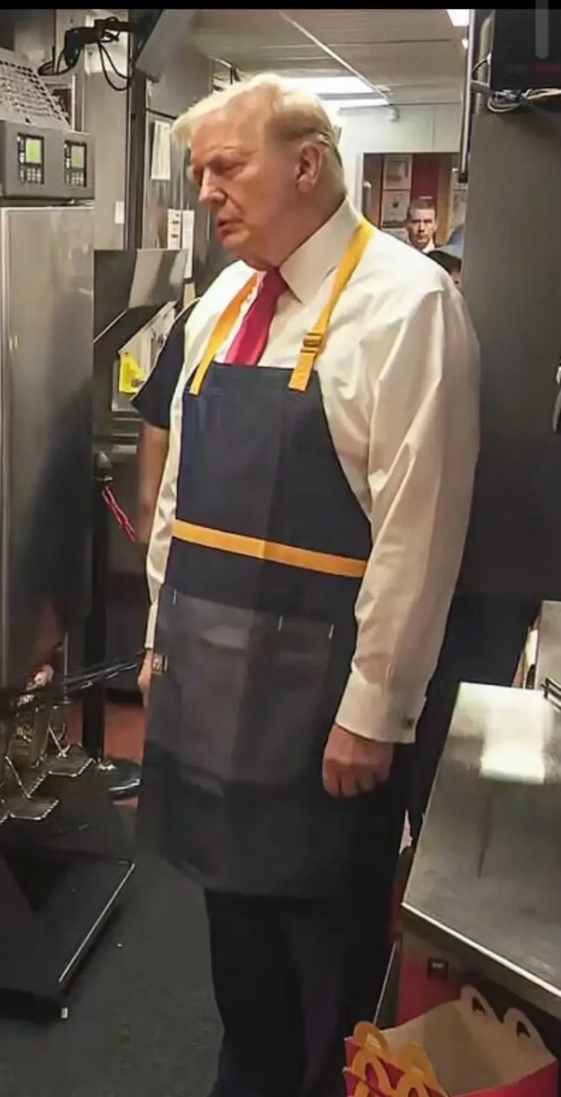 A man with white hair wearing a white dress shirt, red tie, and a dark blue McDonald's apron with yellow straps stands in a…