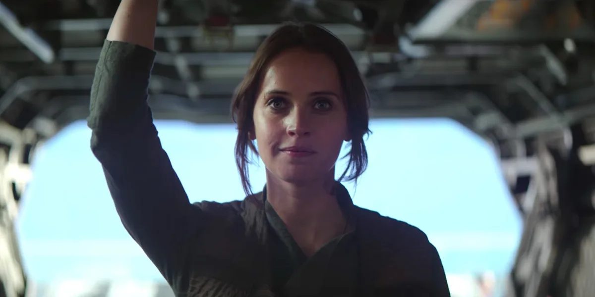 a woman with brown hair and a green shirt raises her right arm in a cockpit-like setting, looking directly at the camera…