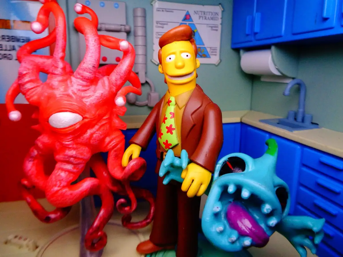 A figurine of the Simpsons character Ned Flanders stands smiling in a miniature kitchen, flanked by two monstrous toy…