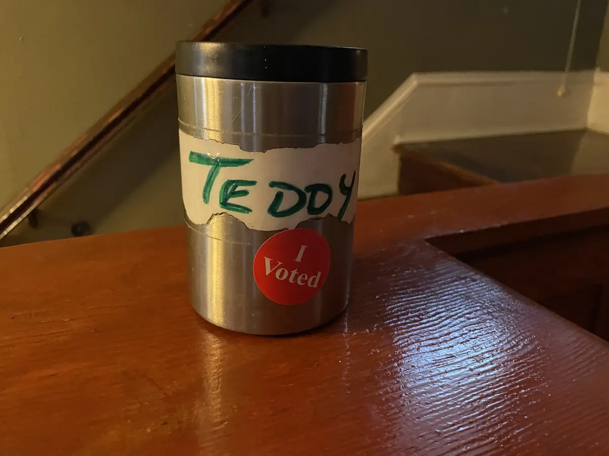 A silver travel mug with a black lid sits on a wooden surface, featuring a torn paper label with 'TEDDY' written in green…