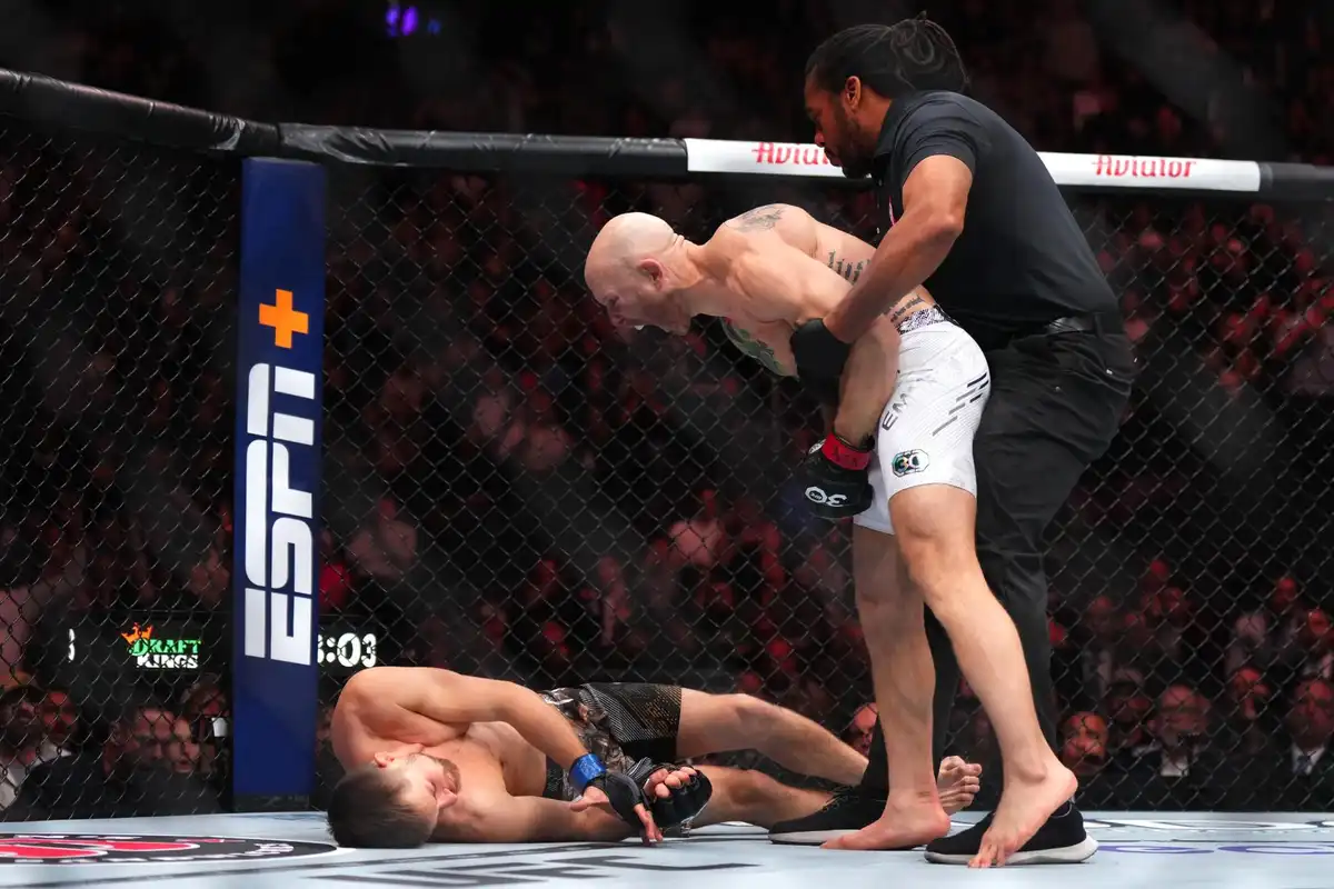 In a UFC octagon, a fighter is being held up by a referee while another fighter lies defeated on the mat. The standing…