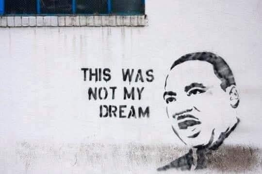A black-and-white stencil graffiti on a white wall depicts a portrait of Martin Luther King Jr. next to the text 'THIS WAS…