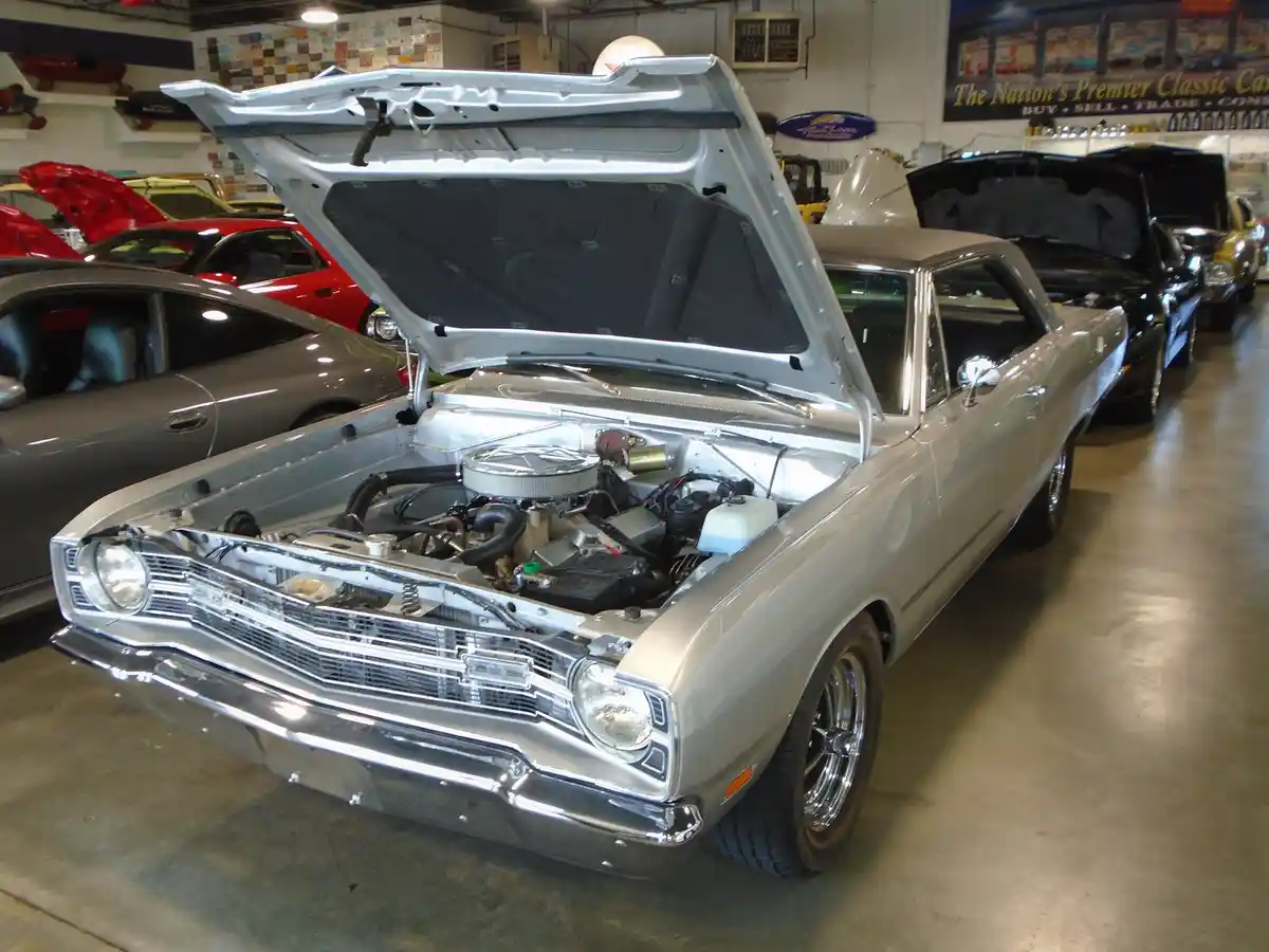 A silver classic American muscle car with its hood open, revealing a detailed engine bay, is the central focus in a well-lit…