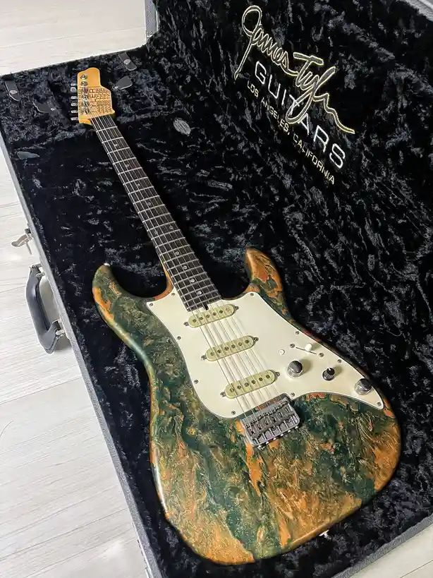 a custom electric guitar with a green and orange swamp-like finish rests in a black velvet-lined case branded 'James Tyler…