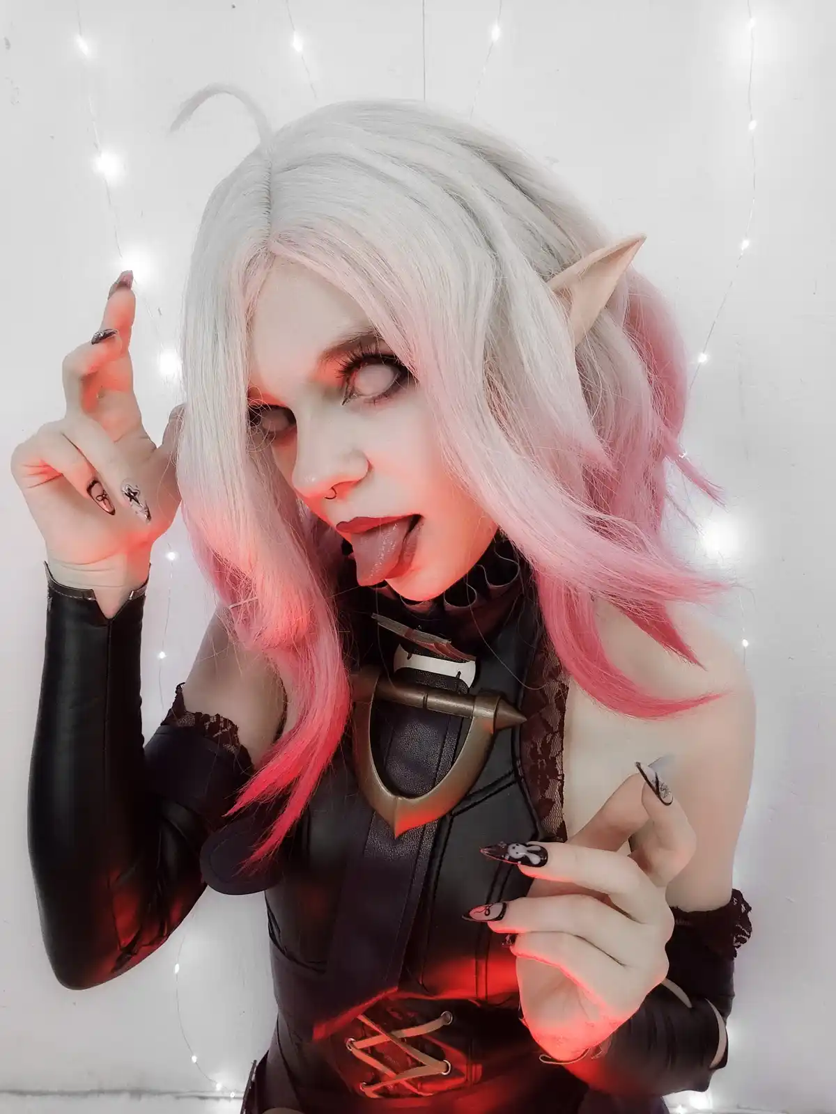 A person in cosplay with white and pink hair, pointed ears, and dark makeup sticks their tongue out playfully while wearing…