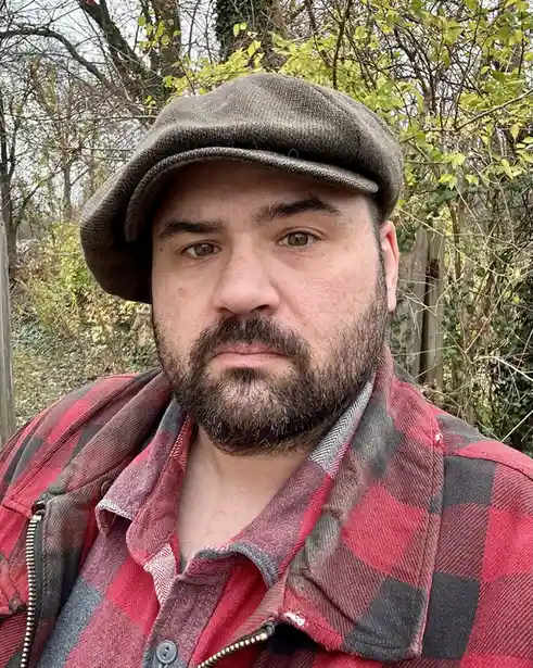 A man with a beard and brown eyes wears a brown flat cap and a red-and-black plaid shirt with a zipper, standing outdoors…