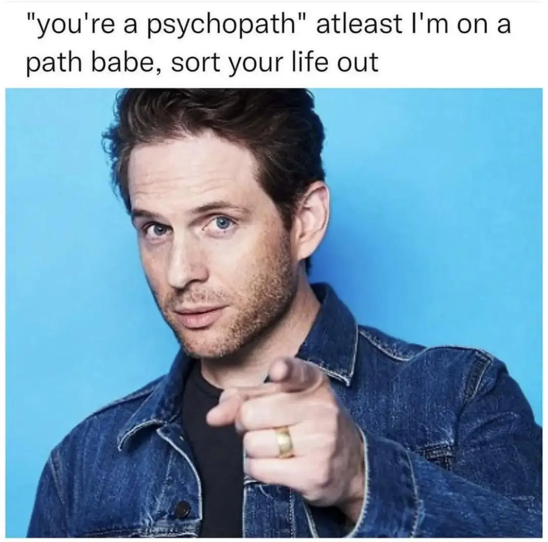 A man with blue eyes and stubble, wearing a denim jacket over a black shirt, points directly at the viewer against a solid…