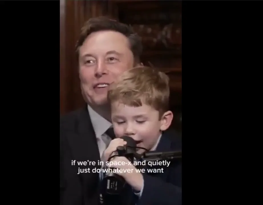 A man and a young boy are both wearing suits, with the boy holding a microphone to his mouth as if singing or speaking. The…