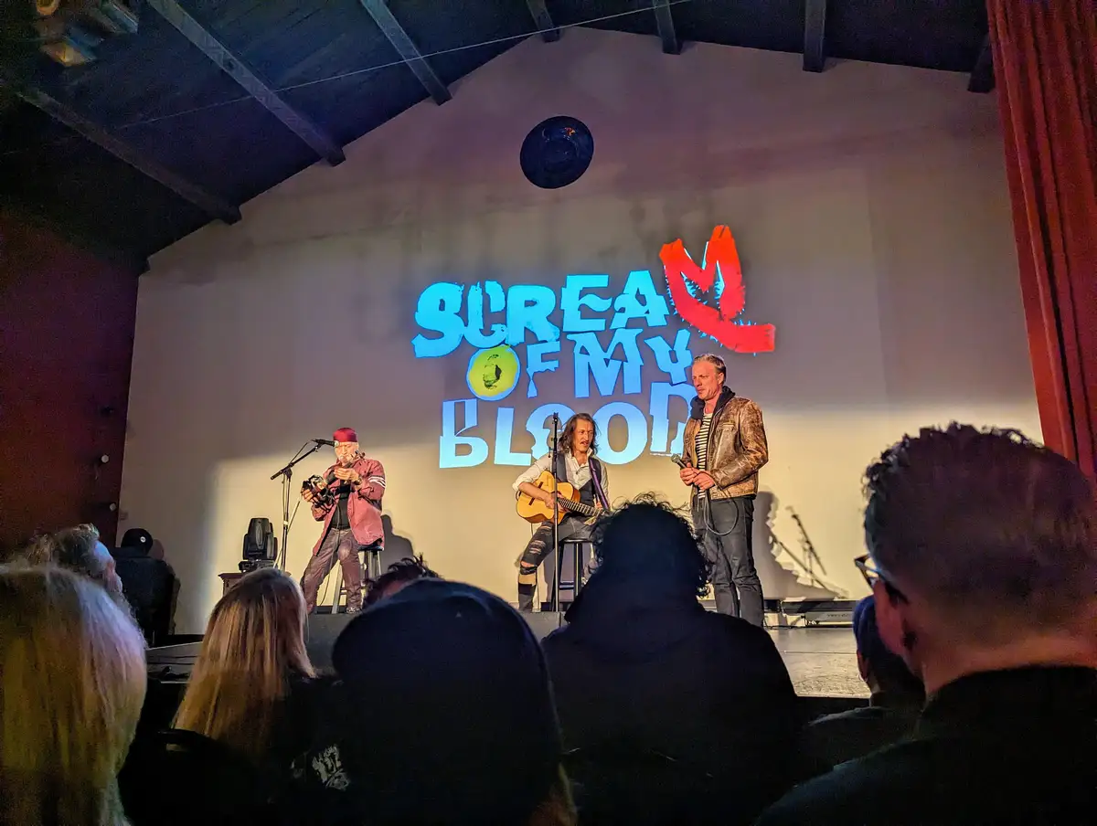 Three musicians perform on a dimly lit stage in front of an audience, with a large projection behind them reading 'SCREAM OF…