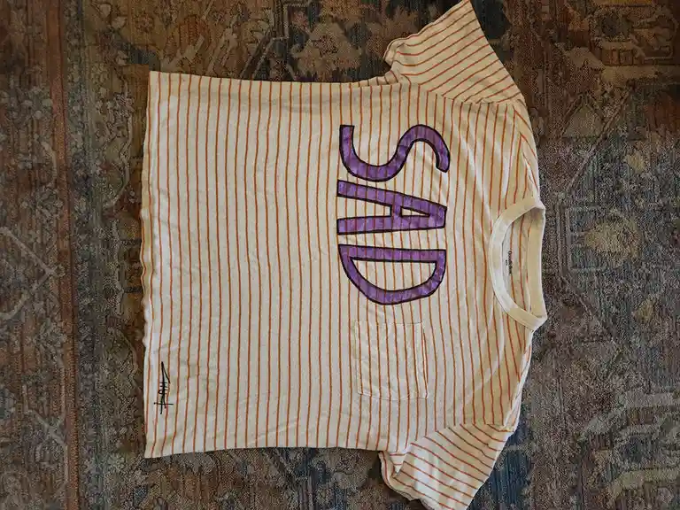 A white t-shirt with thin orange vertical stripes is laid flat on a patterned rug. The word 'SAD' is written vertically in…