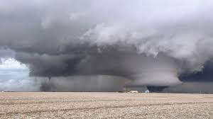 A wide, ominous tornado with a visible funnel cloud dominates a flat, barren landscape under a dark, stormy sky. A small…