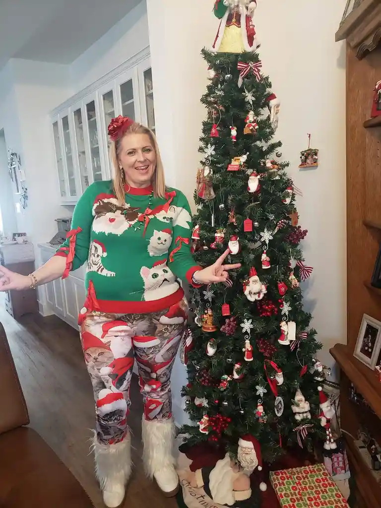 A woman stands beside a heavily decorated Christmas tree, wearing a green sweater and pants both covered in images of cats…