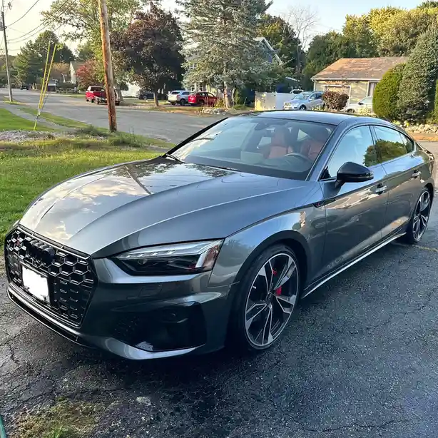 A dark gray Audi sedan is parked on a residential street, its sleek design and sporty wheels visible. The overlaid text…