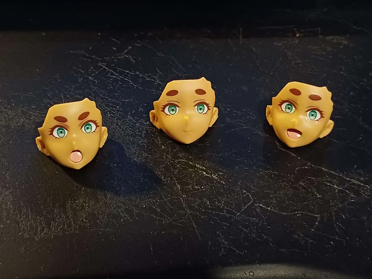 Three anime-style plastic doll heads with green eyes and brown freckles are arranged in a row on a scratched black surface…