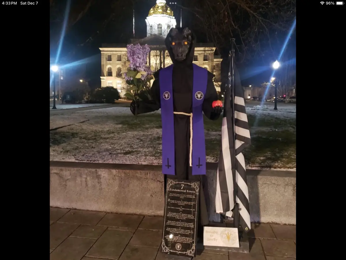 A person in a black hooded costume with a goat head mask and glowing orange eyes stands outdoors at night in front of a lit…