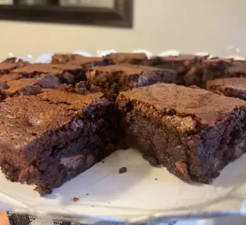 A close-up photo of freshly baked, fudgy brownies cut into squares on a white plate, with one piece slightly separated to…
