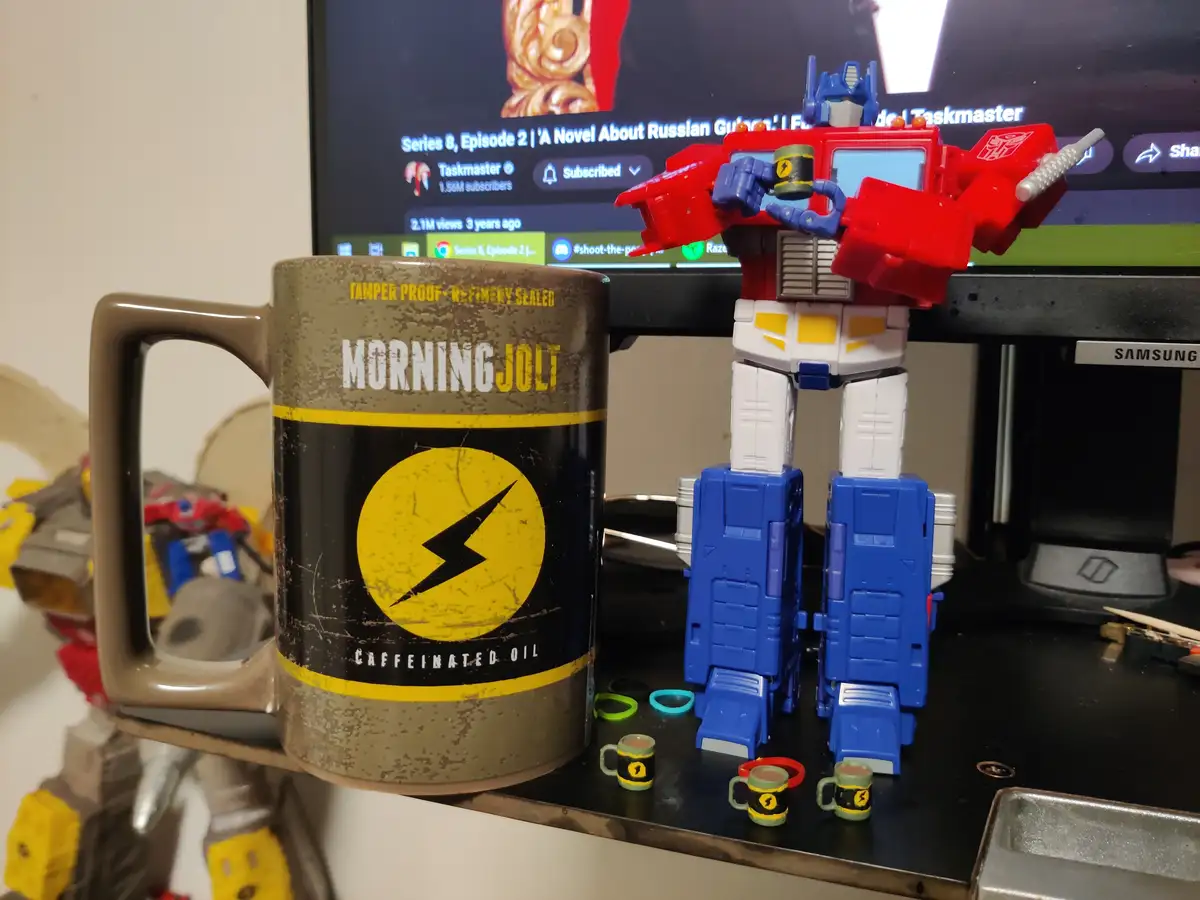 A red, white, and blue Optimus Prime toy robot stands next to a large mug designed to look like a can of 'MorningJolt…