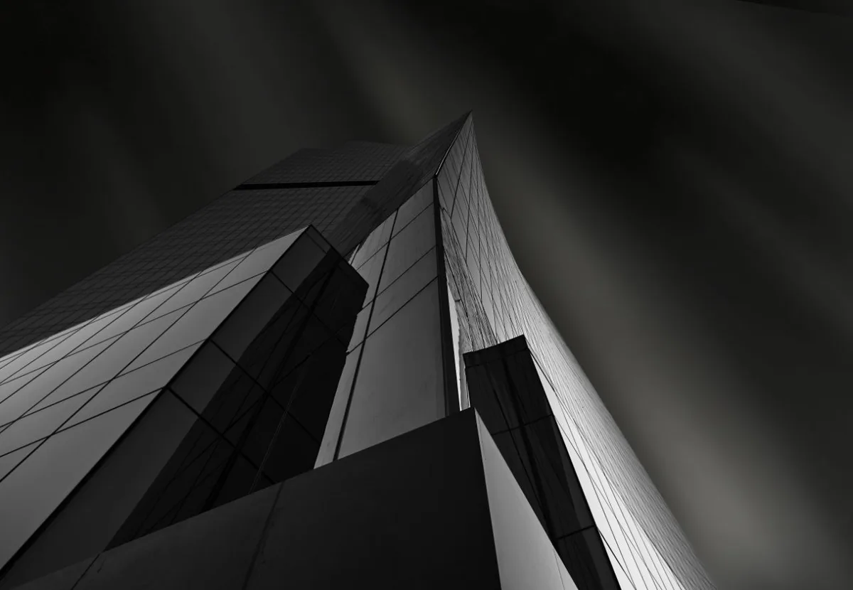 A low-angle black-and-white photograph of a modern skyscraper with sharp geometric lines and reflective glass panels, set…