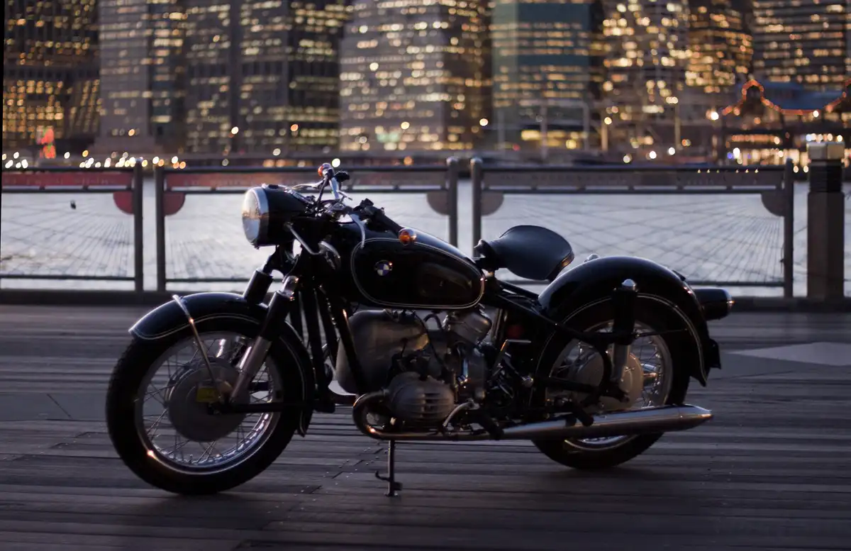 A black vintage BMW motorcycle is parked on a wooden pier at dusk, with a blurred city skyline and glowing lights in the…