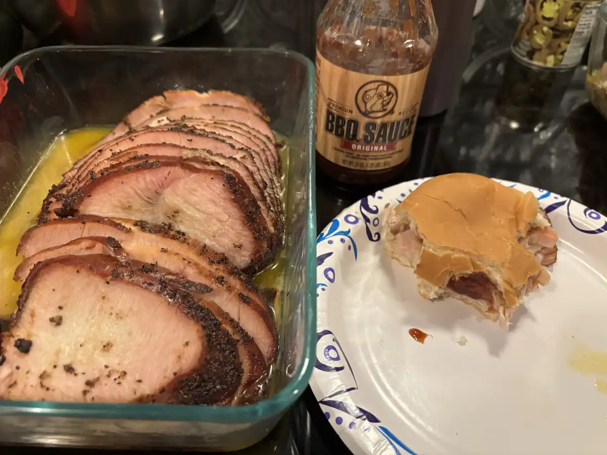 A glass dish holds thinly sliced cooked ham with a dark crust, next to a paper plate with a partially eaten sandwich made…