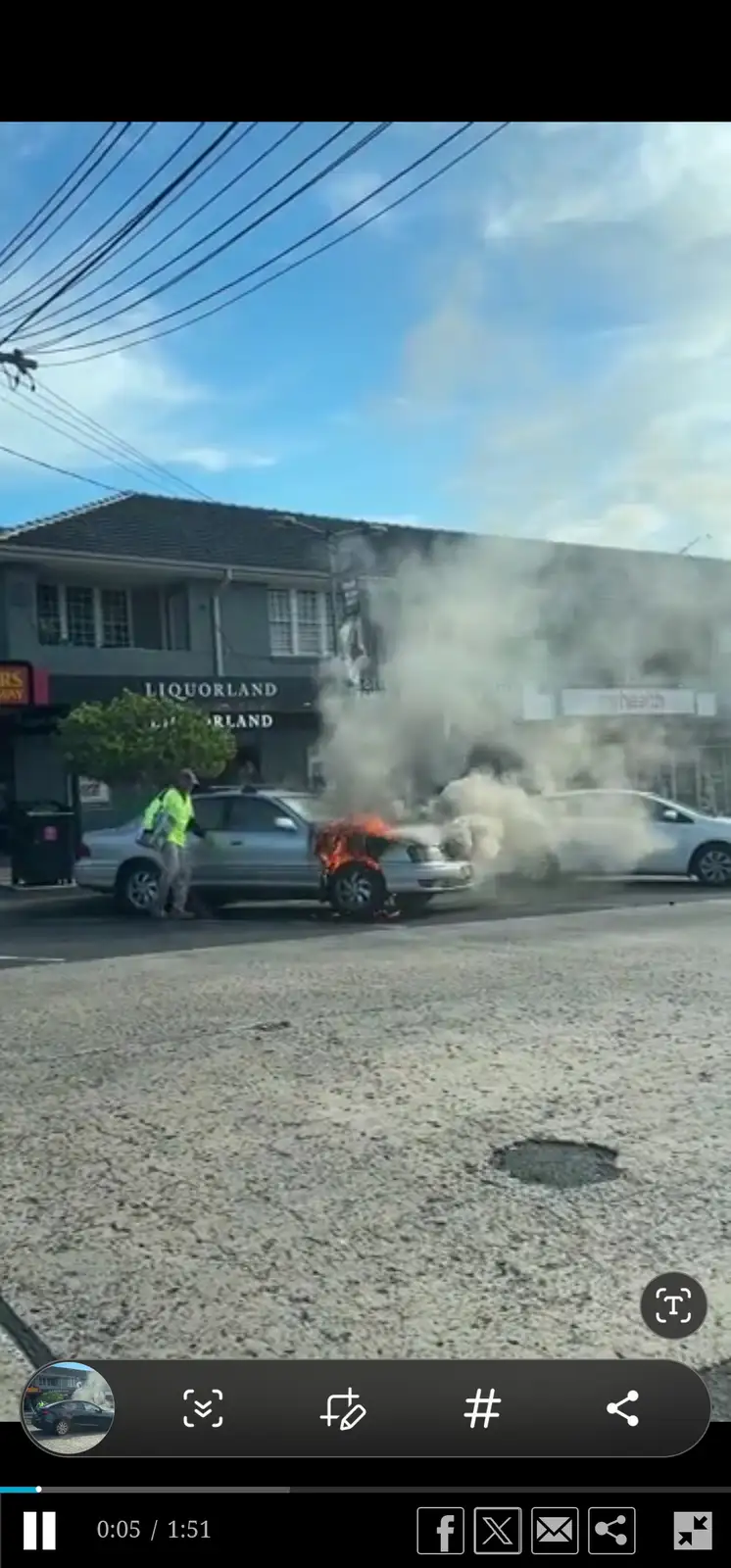 A silver sedan is engulfed in flames and billowing smoke on a street, with a person in a bright yellow safety vest standing…