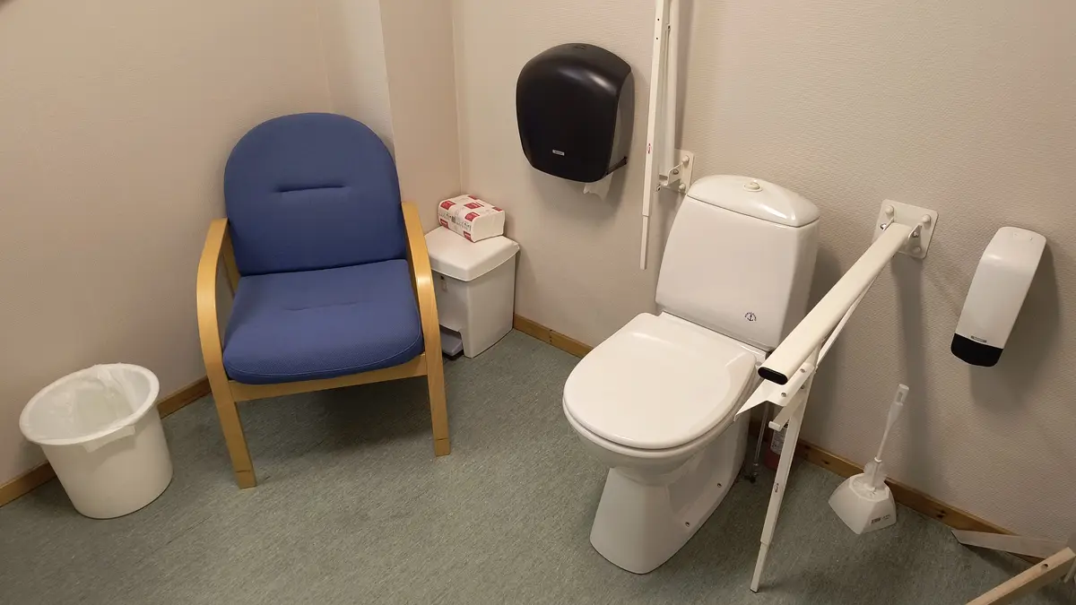 a clean, accessible bathroom features a white toilet with safety rails mounted on the walls, a blue upholstered chair with…