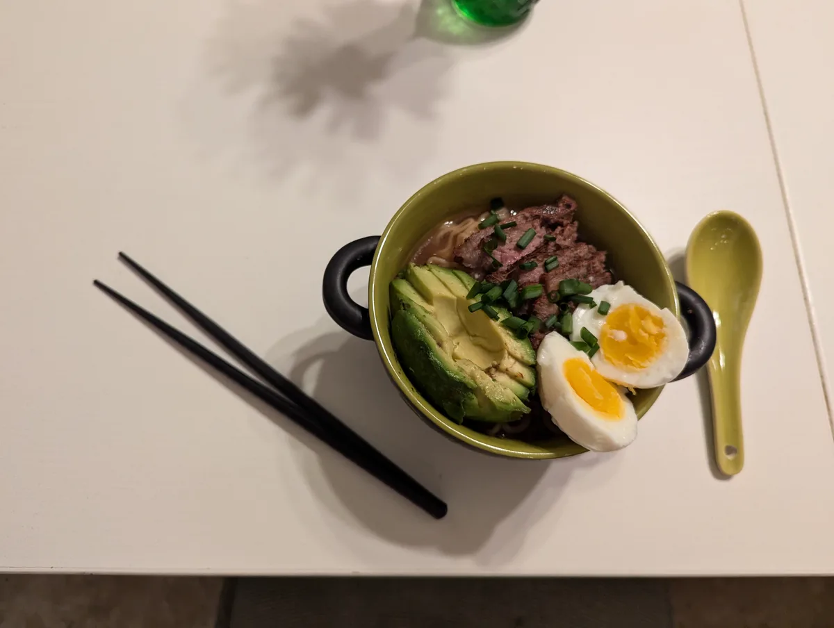 A top-down photo shows a green ceramic bowl with black handles, filled with ramen noodles, sliced avocado, cooked beef…