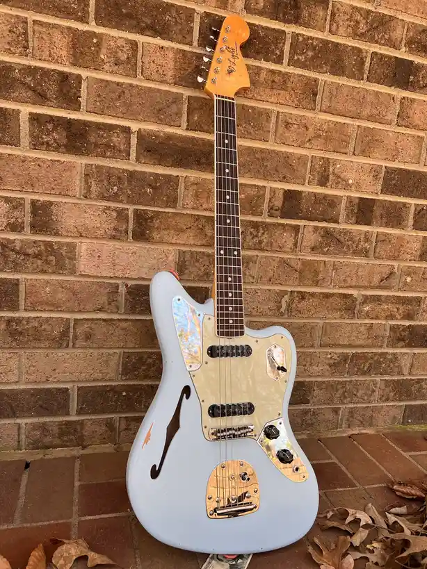 A light blue electric guitar with a cream pickguard and gold hardware leans against a brick wall outdoors, with dry leaves…