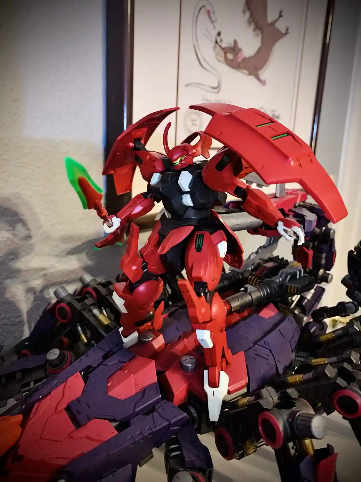 A red and black Gundam model stands atop a pile of disassembled or broken Gundam parts, holding a green and red weapon. The…