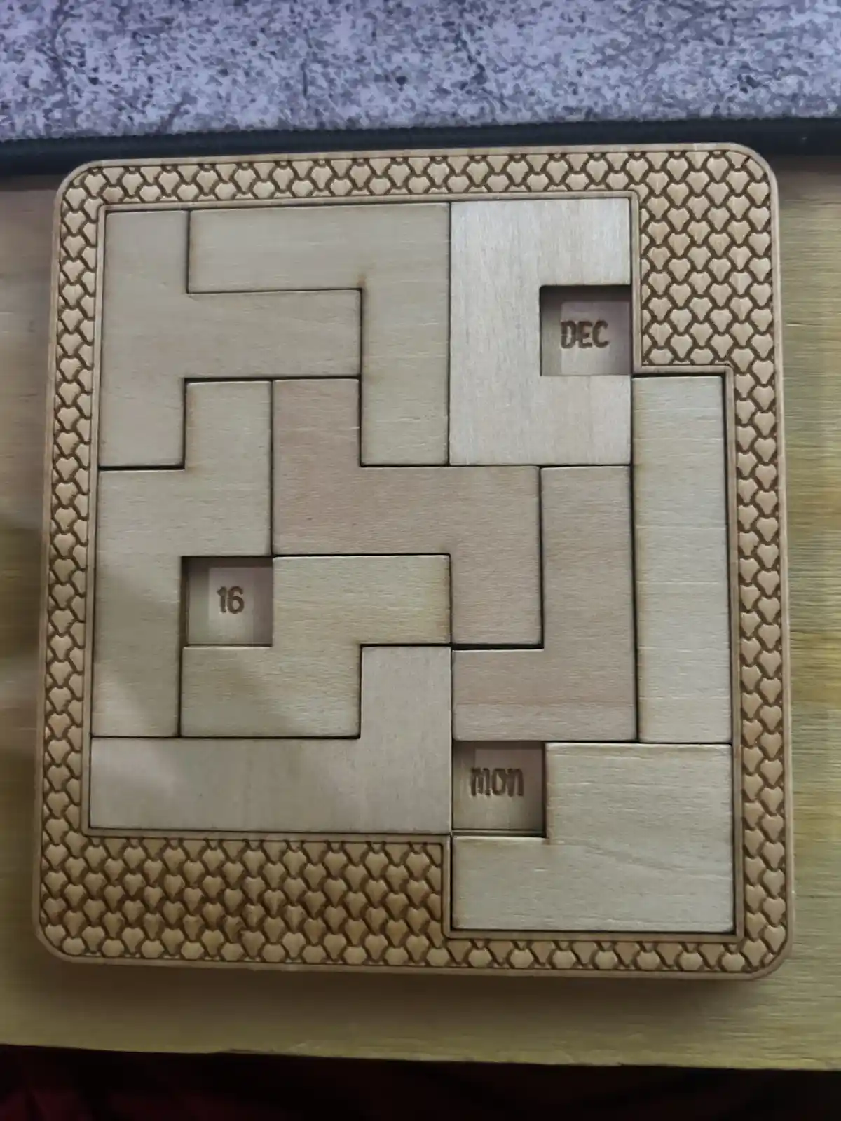 A wooden puzzle calendar with interlocking pieces, showing the date '16' in one recessed square, 'DEC' in another, and 'MON'…
