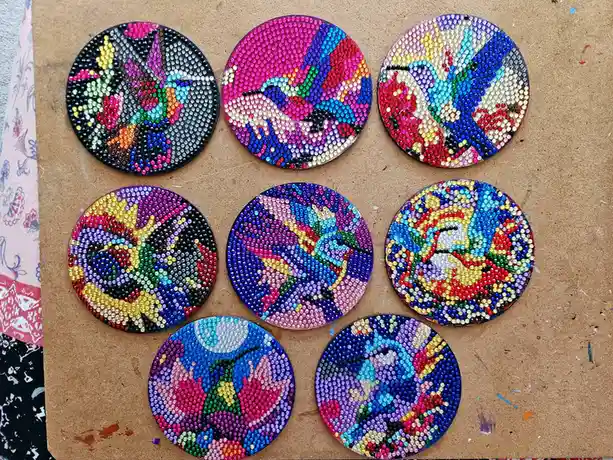 Seven circular artworks made of colorful beads depicting hummingbirds are arranged on a brown cardboard surface. Each piece…