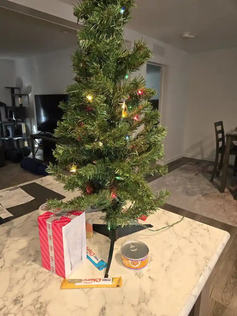 A small, lit artificial Christmas tree stands on a marble kitchen counter, surrounded by everyday items including a T-Mobile…