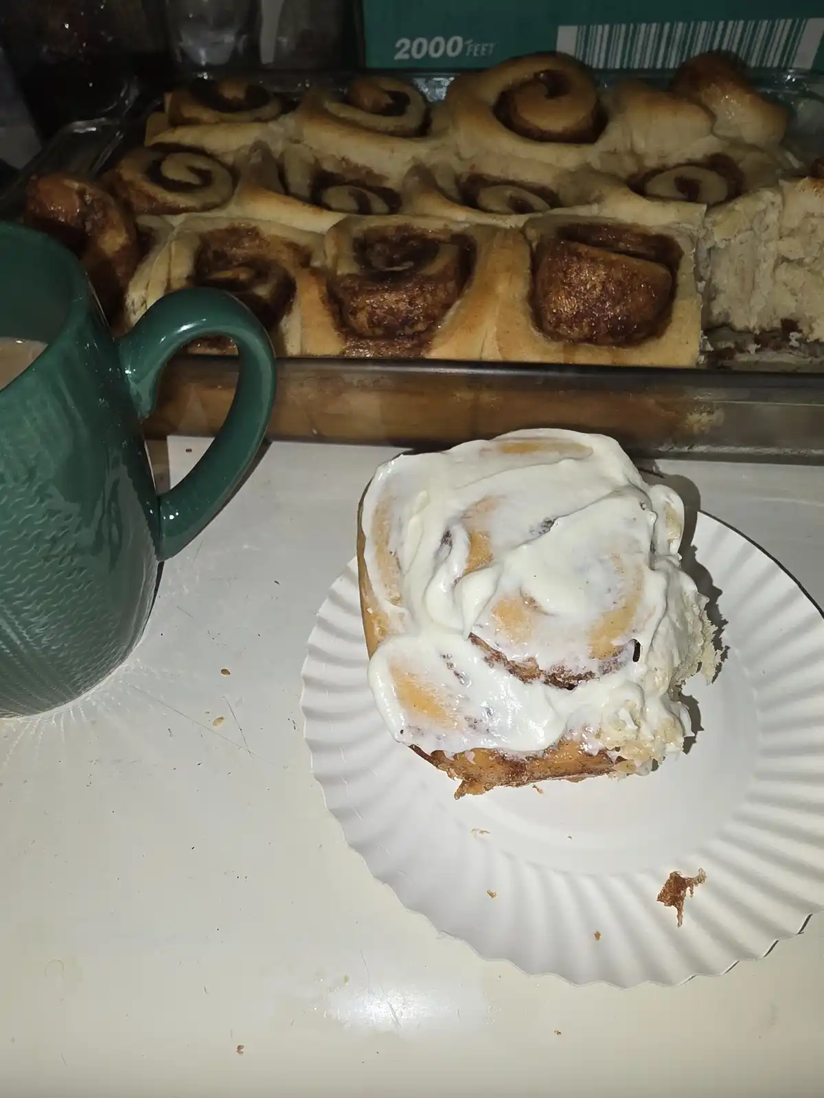 A close-up photo shows a single cinnamon roll with white icing on a paper plate in the foreground, next to a green mug…